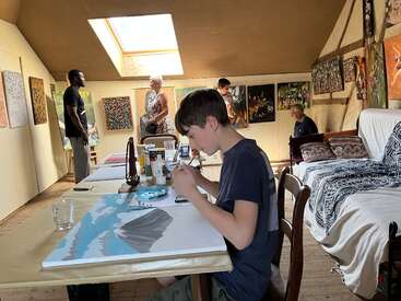 A young boy paints at a table in an art studio, while four adults observe colorful paintings on the walls. Natural light shines through a skylight.