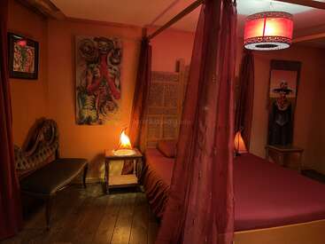 This cozy bedroom features warm lighting, reddish-brown decor, abstract artwork, a canopy bed with drapes, vintage bench, wooden flooring, and a unique, artistic ambiance.