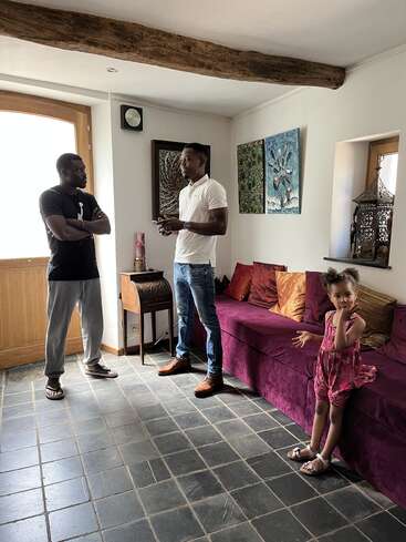 Two men are having a conversation in a cozy, art-filled living room. A young girl in a pink dress stands nearby, curiously observing the scene.