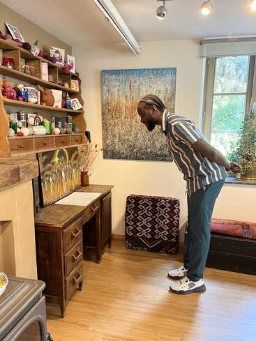 A man in a striped shirt leans over a wooden desk, closely examining artwork in a cozy, decorated room with shelves, paintings, and a window.