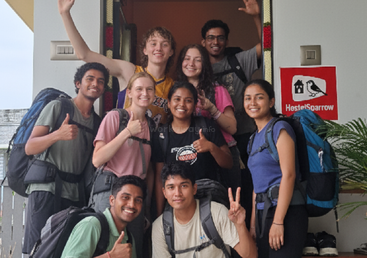 Neun junge Backpacker, fröhlich und lächelnd, posieren gemeinsam am Eingang des Hostel Sparrow. Sie zeigen Friedenszeichen und Daumen nach oben und freuen sich auf ihr Abenteuer.