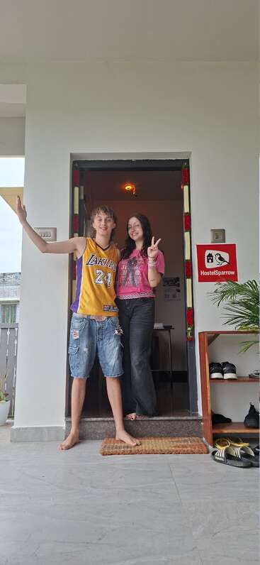 Zwei junge Menschen stehen lächelnd und posierend an einer Tür. Der Junge trägt ein Lakers-Trikot, das Mädchen zeigt ein Friedenszeichen. Das HostelSparrow-Schild ist zu sehen. Entspannte, freundliche Atmosphäre.