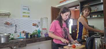Zwei Personen kochen zusammen in einer Küche. Eine Person schneidet Gemüse, während die andere auf dem Herd kocht. Die Küche sieht aufgeräumt aus.