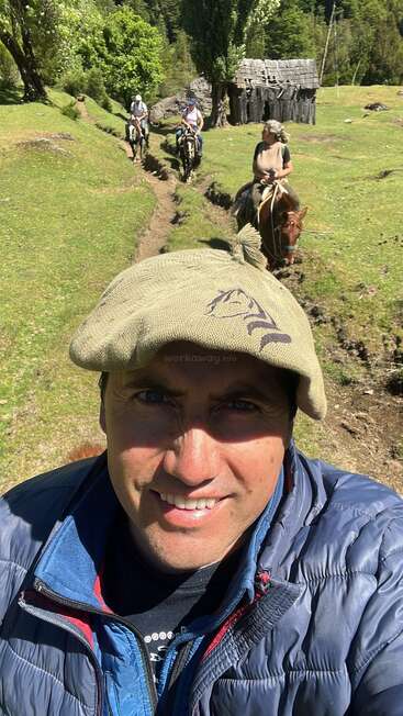 Un hombre con sombrero se toma un selfie al aire libre; detrás de él, tres personas montan a caballo por un sendero cubierto de hierba en el que se ven árboles y una vieja cabaña de madera.