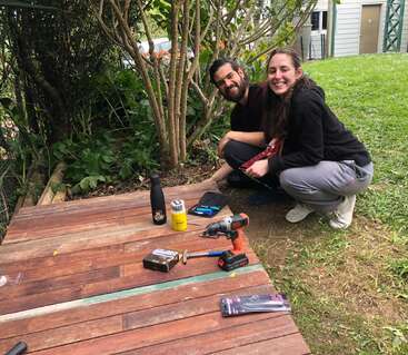 Um homem e uma mulher sorridentes se agacham ao lado de um deck de madeira que estão construindo ao ar livre. Várias ferramentas, bebidas e materiais estão espalhados no deck. Há vegetação ao redor deles.