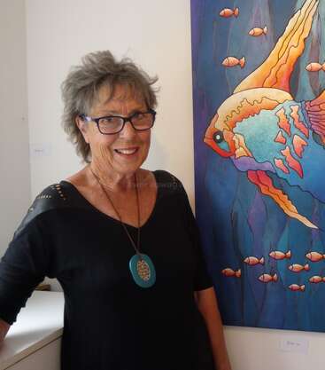 Uma mulher idosa de óculos sorri ao lado de uma pintura de um peixe colorido. Ela usa uma blusa preta e um colar azul, em pé sob a luz brilhante de uma galeria de arte.