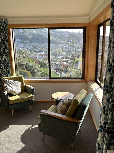 Este acogedor rincón, bañado por la luz del sol, cuenta con dos sillones de estilo retro, una mesita de madera y amplios ventanales que ofrecen unas preciosas vistas a la ladera de un barrio pintoresco.