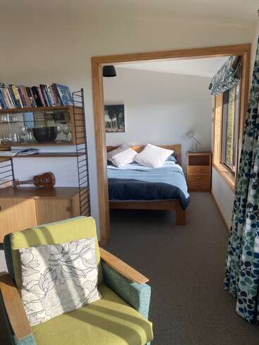 Una acogedora sala con una estantería, un sillón y un ukelele da paso a un luminoso dormitorio con una cama de madera, mesitas de noche y un gran ventanal con cortinas estampadas.
