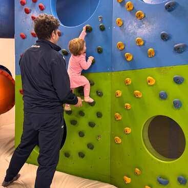 Un hombre observa y ayuda a una niña pequeña vestida de rosa mientras escala un colorido rocódromo interior, con agarres azules, verdes y amarillos.