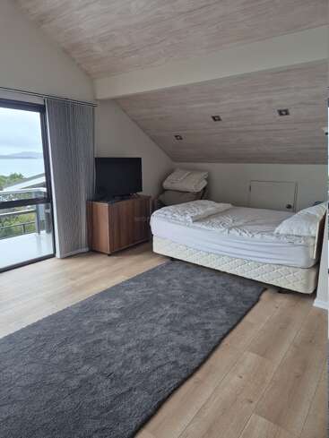 Une chambre mansardée confortable avec un plafond en bois incliné, un lit défait, une télévision sur un support en bois, une grande fenêtre avec une vue panoramique et un long tapis gris.