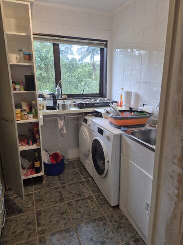 Une petite buanderie fonctionnelle avec un lave-linge, un sèche-linge, un évier, des étagères ouvertes, des ustensiles de cuisine, un sol carrelé et une fenêtre donnant sur la verdure.