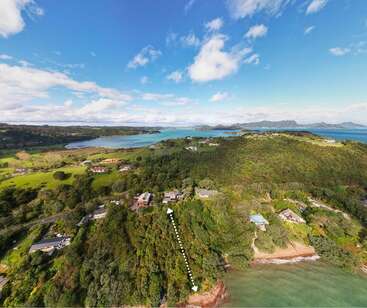 Cette image aérienne montre un paysage côtier verdoyant avec des maisons dispersées, des collines ondulantes, un ciel bleu clair, des baies d'eau et des îles montagneuses lointaines. Serein et pittoresque.