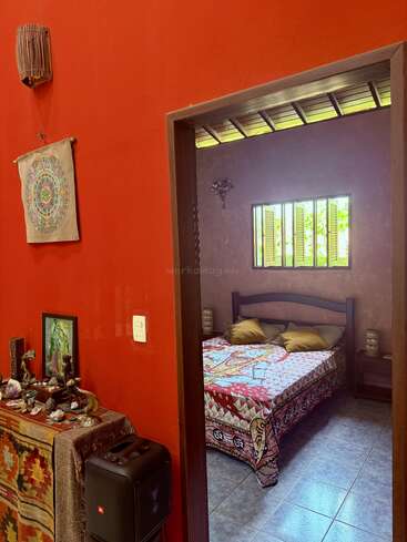 El interior de una casa acogedora y colorida, con paredes rojas, adornos espirituales, una colcha ornamentada, contraventanas abiertas, suelo de baldosas y un pequeño altar con cristales y figuritas.