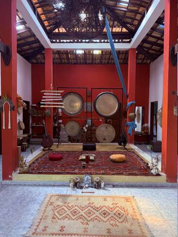 Esta imagen muestra una habitación serena, de techo abierto y paredes rojas, con gongs, instrumentos musicales, alfombras de colores, cojines y estatuas espirituales, que crean una atmósfera de calma y meditación.