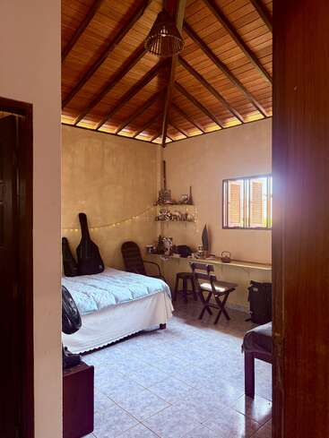 Un acogedor dormitorio con un alto techo de madera, cama, escritorio, silla, fundas de guitarra, luces de cuerda, suelo de baldosas y luz solar que entra por una ventana abierta.