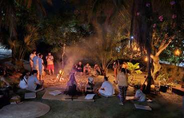 Un grupo de personas se reúne por la noche en torno a una cálida hoguera, rodeados de plantas tropicales, compartiendo historias, relajándose en colchonetas y disfrutando de un acogedor ambiente al aire libre.