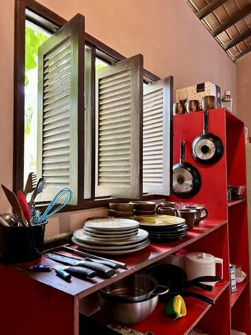 Un acogedor rincón de cocina cuenta con estanterías rojas llenas de platos, tazas, utensilios, ollas y sartenes cuidadosamente apilados. La luz del sol entra a raudales por las ventanas con contraventanas de madera.