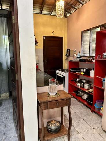 Esta acogedora cocina cuenta con una mesa de madera con una jarra de cristal, estanterías rojas abiertas llenas de utensilios de cocina, un horno, suelos de baldosas y una lámpara de techo rústica.