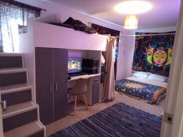 Une chambre confortable et colorée avec un lit en mezzanine au-dessus d'un bureau et d'une armoire, un lit double, une tapisserie murale vibrante, des marches et un tapis bleu rayé.