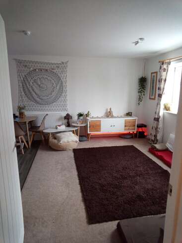 Une pièce confortable et minimaliste avec une moquette beige, un tapis brun foncé, une table basse, des chaises, des plantes, une tapisserie murale, une statue de Bouddha, une fenêtre avec des rideaux floraux et un éclairage doux.