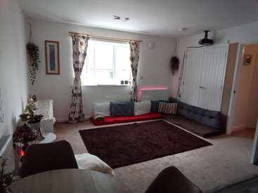 Un salon confortable et minimaliste avec un grand tapis marron, des coussins bas, une fenêtre avec des rideaux floraux, des plantes, des objets décoratifs et un projecteur fixé au plafond.