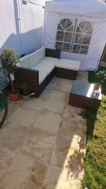 Un patio confortable avec des meubles en osier, des coussins crème, des plantes en pot, un gazebo blanc, un sol carrelé, une table basse, des tasses, un tuyau d'arrosage, une roue de vélo et un peu de soleil.