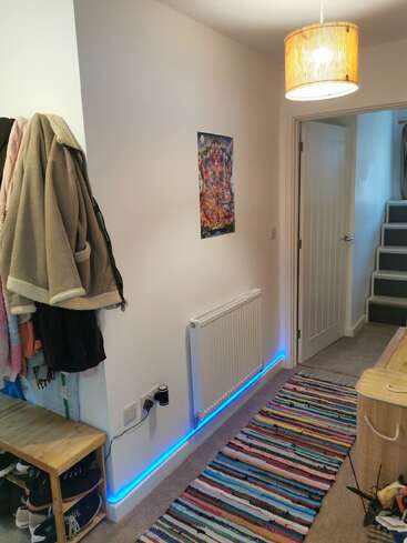 Un couloir confortable avec un tapis rayé, des manteaux colorés sur une étagère, un banc en bois, des œuvres d'art murales, des lampes LED bleues et des escaliers menant vers le haut.