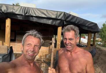 Two shirtless men smiling, pose outdoors in front of a partially constructed wooden structure. One holds a tool, both appear to be working on a building project.