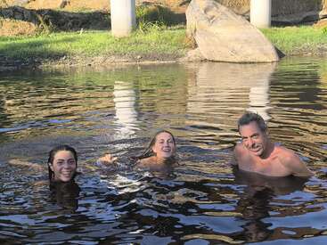 Three people are swimming and enjoying themselves in a calm, natural outdoor body of water, with sunlight and green grass visible in the background.