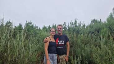 A man and woman stand closely together in front of green bushes and trees. The man wears a "Free Palestine" shirt, while the woman wears glasses.