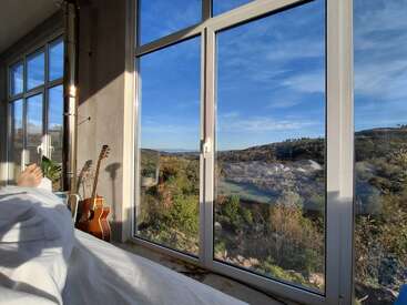 A cozy bed beside large windows overlooks a beautiful scenic view of green hills under a clear blue sky. A guitar and plant add warmth.