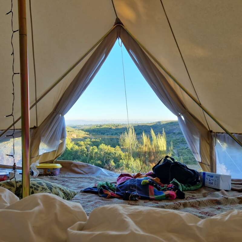 The image shows the interior of a tent with a wooden frame, featuring a table and chairs, set against a backdrop of a grassy field and a tree line. The tent is surrounded by a fence.