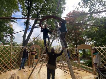 Ein paar Leute bauen draußen zusammen das Holzgerüst einer Jurte auf. Zwei von ihnen stehen auf Leitern und montieren den zentralen Dachring unter Bäumen.