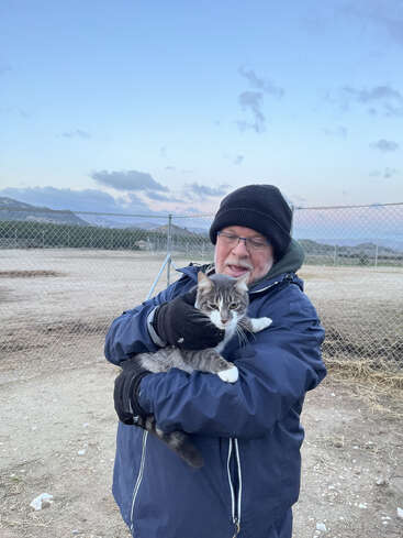 A man bundled in a blue jacket, gloves, and black beanie lovingly holds a gray and white cat outdoors, with mountains and a chain-link fence behind them.