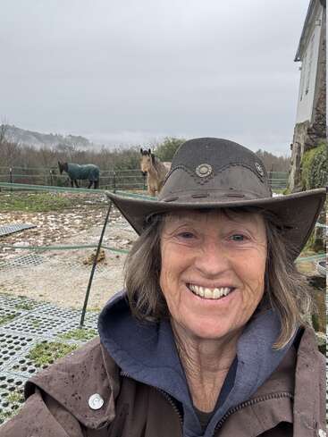 Uma mulher sorridente, com uma capa de chuva marrom e chapéu, tira uma selfie ao ar livre. Atrás dela, tem dois cavalos, um deles coberto, num dia nublado e nebuloso no campo.