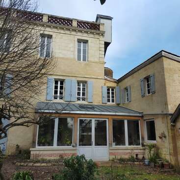 This image shows a charming old stone house with blue shutters, a glass-enclosed porch, potted plants, bare trees, and a small garden under a clear sky.