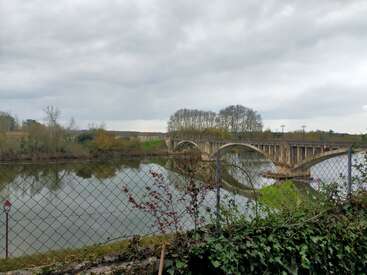 Um dia nublado mostra uma ponte de pedra em arco cruzando um rio calmo, cercado por árvores. A hera e um primeiro plano cercado criam uma atmosfera natural e tranquila.
