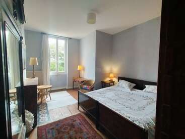 Um quarto aconchegante e bem iluminado, com uma janela grande, piso de madeira, cama de casal, abajures, poltronas, uma mesinha redonda, guarda-roupa e uma decoração simples e convidativa.