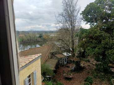A vista da janela mostra uma casa amarela, um jardim com árvores, uma rede e um rio. O céu nublado e a paisagem distante completam a cena rural tranquila.