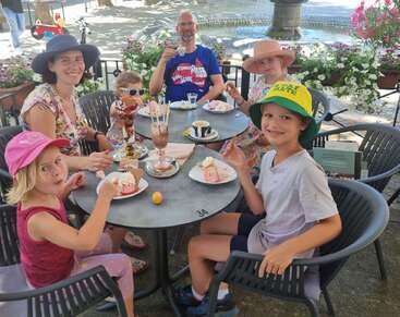 A family of five enjoys ice cream and coffee at an outdoor café, smiling together at a round table surrounded by flowers and a sunny atmosphere.