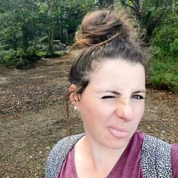 Une femme aux cheveux en chignon désordonné se tient à l'extérieur, dans une zone boisée, faisant un clin d'œil et tirant la langue de manière ludique, vêtue d'une chemise marron et d'un sac à dos.