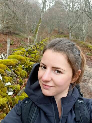 Une femme vêtue d'une veste sombre prend un selfie en plein air. Derrière elle, des rochers couverts de mousse et des arbres dénudés créent une scène forestière naturelle et paisible en automne.