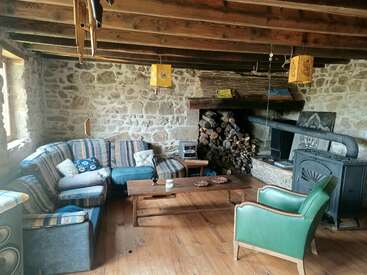 Ce salon rustique et confortable présente des murs en pierre, des poutres en bois apparentes, un poêle à bois, du bois de chauffage empilé, un canapé sectionnel rayé, un fauteuil vert et des éléments de décoration en bois.