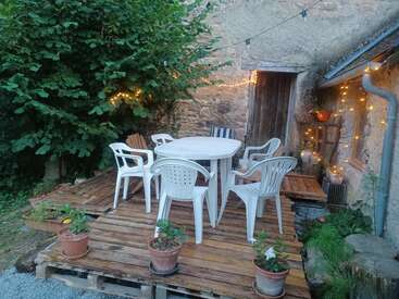 Ce patio rustique se compose d'une terrasse en palettes de bois, de chaises en plastique blanc, d'une table ronde, de plantes en pot, de guirlandes électriques, de verdure et d'un mur de pierre en toile de fond. Ambiance chaleureuse.