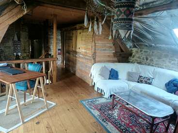 Cette chambre mansardée confortable est dotée de murs en bois, d'un canapé sectionnel blanc, de coussins colorés, d'une table basse en verre, d'un bureau avec un ordinateur portable et d'une décoration rustique et bohémienne.
