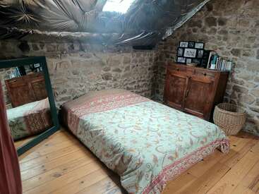 Cette chambre mansardée confortable comprend un lit bas avec une couverture à motifs, des planchers en bois, des murs en pierre, un grand miroir, une armoire rustique, des livres, un panier et une lucarne.