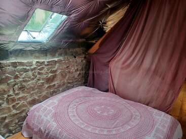 Cette chambre mansardée confortable est dotée d'un mur en pierre, d'un plafond incliné avec une lucarne et d'un lit recouvert d'une couverture à motifs roses sous un tissu drapé pour plus d'intimité.