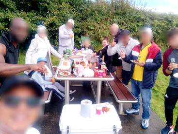 Un groupe de personnes profite d'un pique-nique en plein air, réunies autour d'une table avec de la nourriture, des boissons et des en-cas. Sourires, rires et ambiance joyeuse sont au rendez-vous.