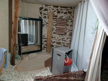 Cette salle de bains rustique présente un mur en pierre, un grand miroir, une baignoire triangulaire, un tapis, des poutres en bois, des rideaux et un radiateur ou un appareil gris avec un objet rouge.