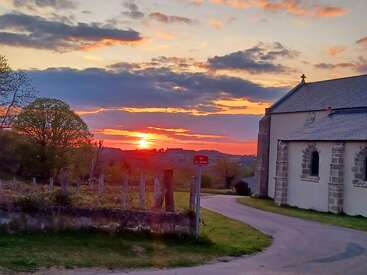 Un coucher de soleil vibrant jette des couleurs chaudes dans le ciel. Une route rurale serpente à côté d'une vieille église, des champs verts, des arbres et une clôture en bois rustique complètent cette scène paisible.